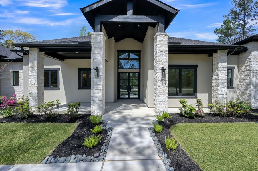 A stunning architectural statement welcomes you home—featuring a dramatic covered entry framed by natural stone columns, bold rooflines, and sleek black accents, all complemented by curated landscaping and a serene approach.