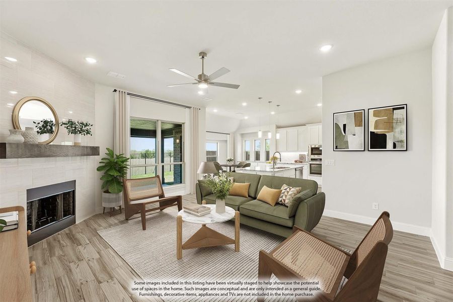Open-concept living room with white fireplace, ceiling fan, hardwood floors, and kitchen visible in background