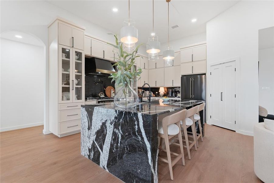 Gourmet kitchen featuring a waterfall island with a dark stone countertop, light wood-finish flooring, recessed lighting, and glass-front upper cabinetry