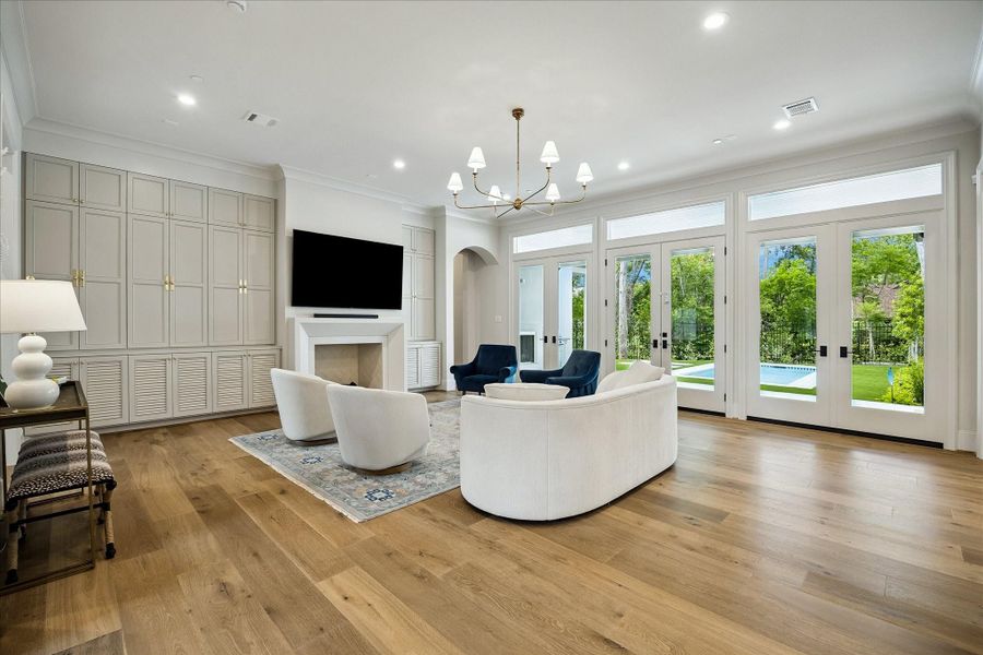 The living room unfolds with refined elegance, showcasing French white oak wide plank hardwood flooring, soaring 11-foot ceilings, custom trim and crown molding. A wood-burning fireplace with surrounding built-ins anchors the space, while a wall of French doors frames serene views of the backyard and fills the room with natural light.