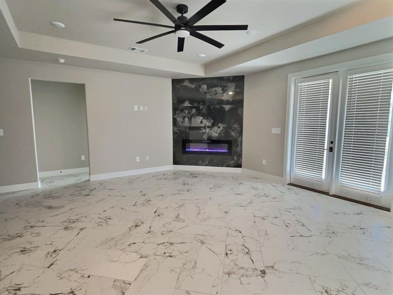 Unfurnished living room featuring a glass covered fireplace, a raised ceiling, light marble finish floors, recessed lighting, and ceiling fan