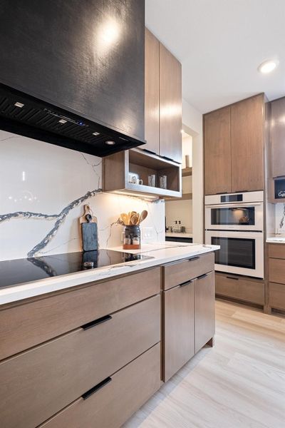 Kitchen with range hood, open shelves, modern cabinets, multiple ovens, and black electric stovetop