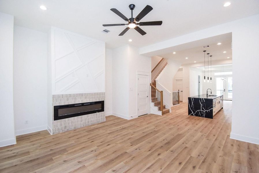 This photo showcases a modern, open-concept living space with light wood flooring, a sleek electric fireplace, and a stylish kitchen island with black marble countertops. The area is bright, featuring recessed lighting and a ceiling fan, with a staircase leading to the upper level.