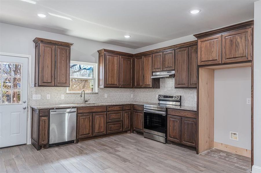 Kitchen featuring light wood-type flooring, under cabinet range hood, light stone counters, and stainless steel appliances