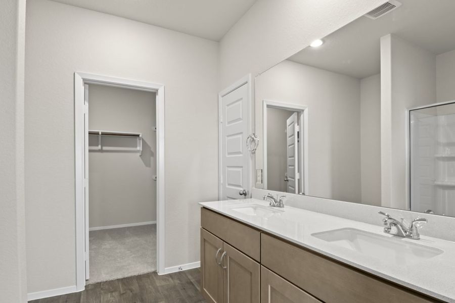 Image of a primary bedroom with light brown cabinets, a double jack and jill white vanity, a large window and a walk-in closet