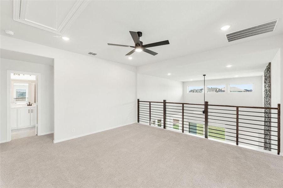 Open upstairs loft with carpet, ceiling fan, metal railing overlooking lower level, and adjacent bathroom doorway