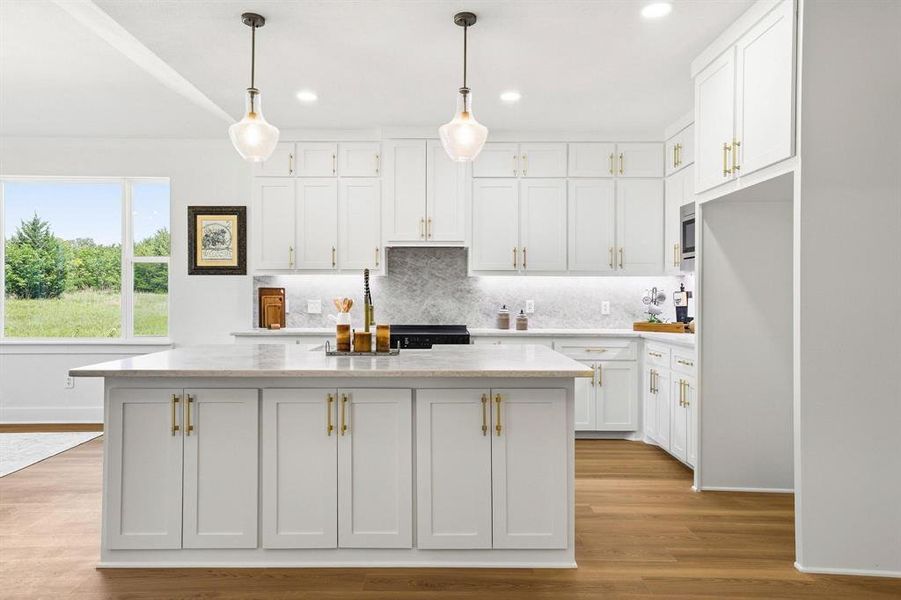 Contemporary kitchen featuring a large central island with a light-toned countertop, white cabinetry with gold-toned hardware, a full-height backsplash, wood-finish flooring, and pendant lighting Contemporary kitchen featuring a large central island with a light-toned countertop, white cabinetry with gold-toned hardware, a full-height backsplash, wood-finish flooring, and pendant lighting