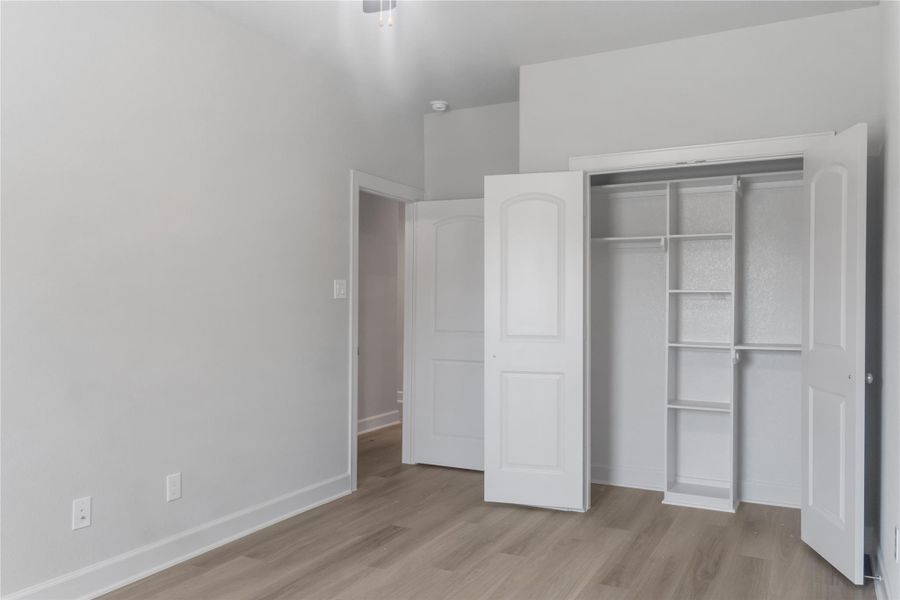 This photo shows a bright, modern room with light-colored walls and wood flooring. It features an open, spacious closet with built-in shelves and double doors. The room is well-lit with natural light, offering a clean and inviting space.