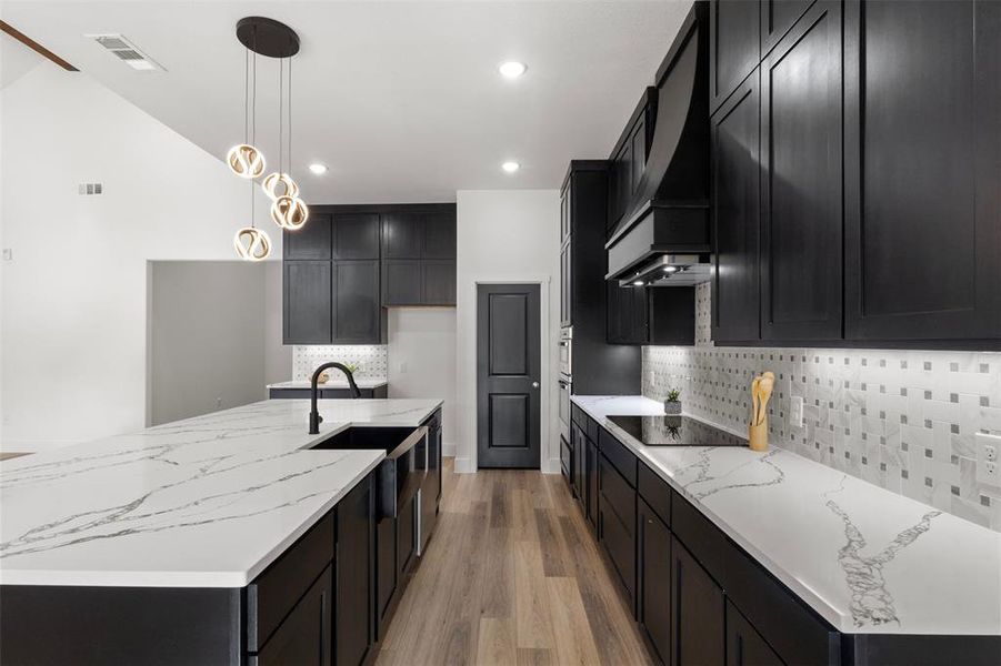 Kitchen with dark cabinetry, light stone counters, pendant lighting, an island with sink, and light wood-style flooring