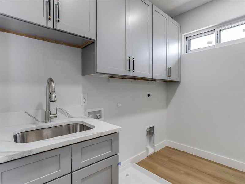 Laundry room featuring light gray cabinetry with black and brass hardware, a stainless steel undermount sink, a white countertop, and wood-finish flooring Laundry room featuring light gray cabinetry with black and brass hardware, a stainless steel undermount sink, a white countertop, and wood-finish flooring
