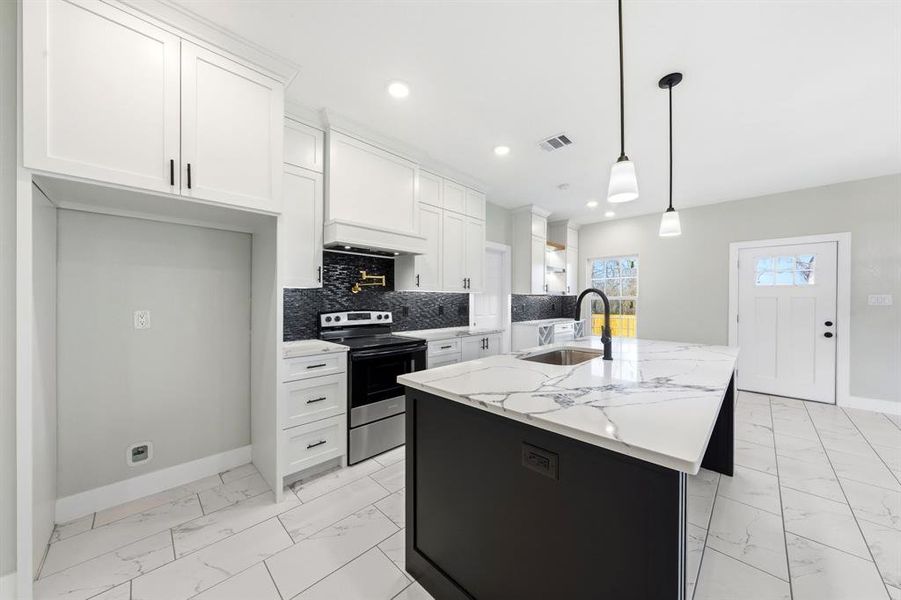 Kitchen with electric stove, a kitchen island with sink, light stone counters, light marble finish flooring, and hanging light fixtures