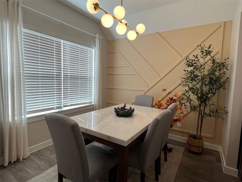 This dining area features a modern design elegant accent wall. Large windows with blinds provide ample natural light, complemented by a stylish overhead light fixture