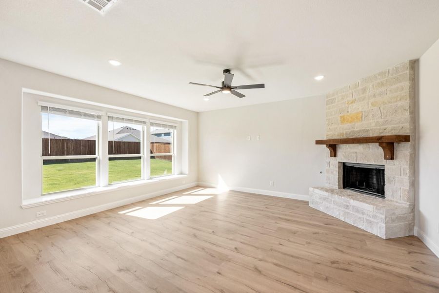 Open living room with stone fireplace, wood mantel, hardwood floors, and large windows overlooking backyard