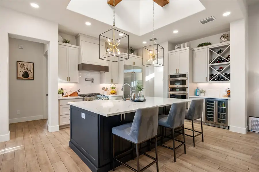 Kitchen with decorative light fixtures, white cabinetry, an island with sink, wine cooler, and light stone counters Kitchen with decorative light fixtures, white cabinetry, an island with sink, wine cooler, and light stone counters