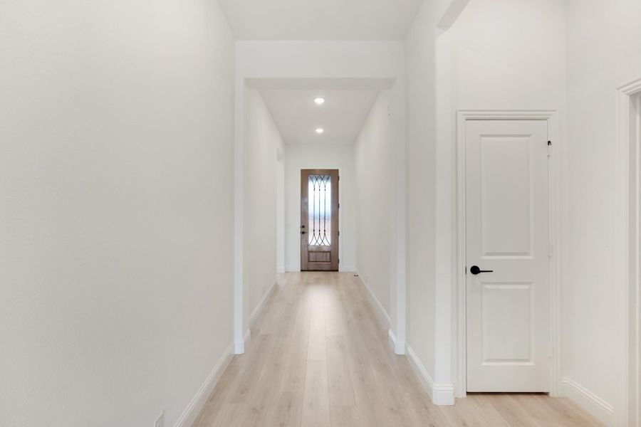 Bright hallway with light wood floors, white walls, and a wood front door with glass panel at the end