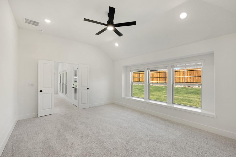 Empty bedroom with vaulted ceiling, black ceiling fan, carpet flooring, and large windows overlooking a fenced backyard