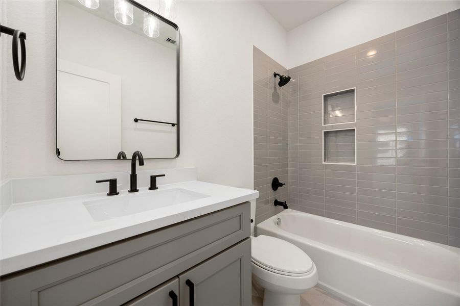This secondary bathroom boasts tile flooring, a bath/shower combo with a stylish tile surround, stained wood cabinetry, elegant light countertops, a mirror, and sleek fixtures complemented by modern finishes.