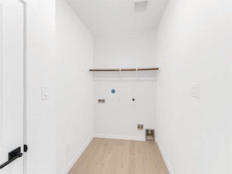 Laundry room with light wood flooring, white walls, and hookups for a washer and dryer. There's a white door with a black handle and a shelf with a hanging rod above the hookups.