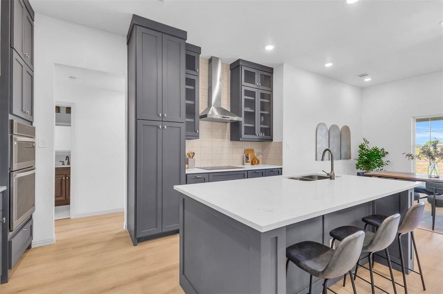 Kitchen featuring decorative backsplash, glass insert cabinets, light wood-type flooring, wall chimney exhaust hood, and gray cabinetry Kitchen featuring decorative backsplash, glass insert cabinets, light wood-type flooring, wall chimney exhaust hood, and gray cabinetry
