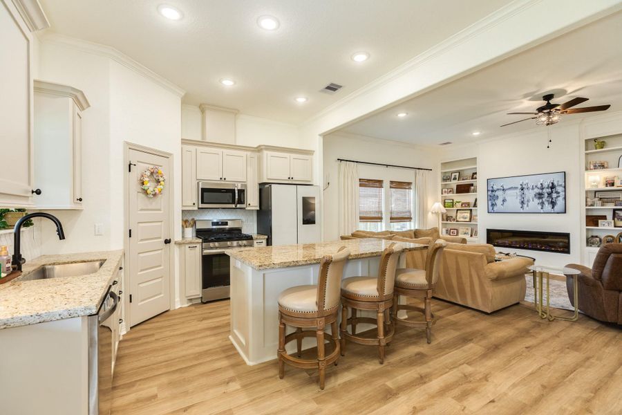 This inviting open-concept space features a modern kitchen with granite countertops, stainless steel appliances, and a central island with seating. It seamlessly connects to a cozy living area with built-in shelves, a fireplace, and large windows, creating a bright and welcoming atmosphere.