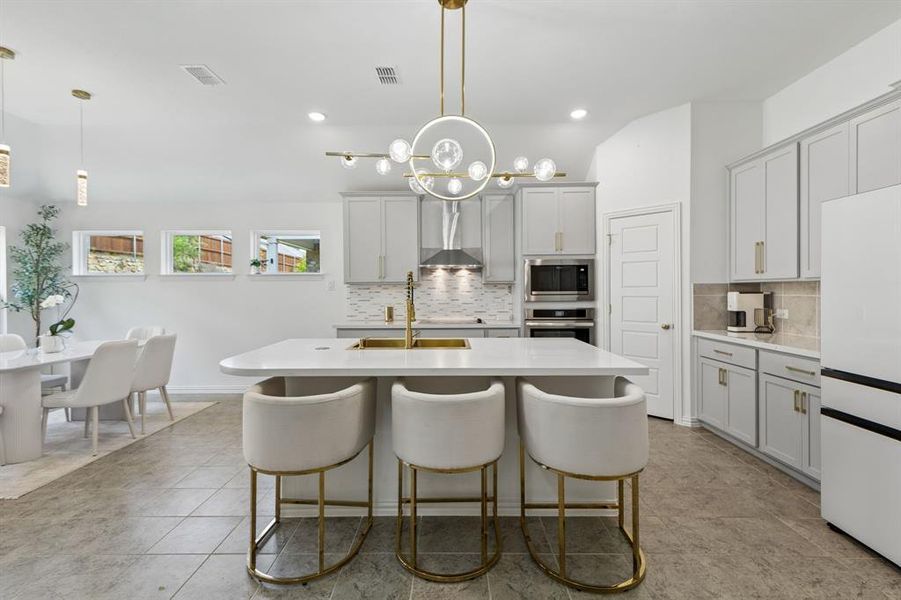 Contemporary kitchen featuring a large central island with seating, light gray cabinetry, stainless steel appliances, and a decorative overhead light fixture