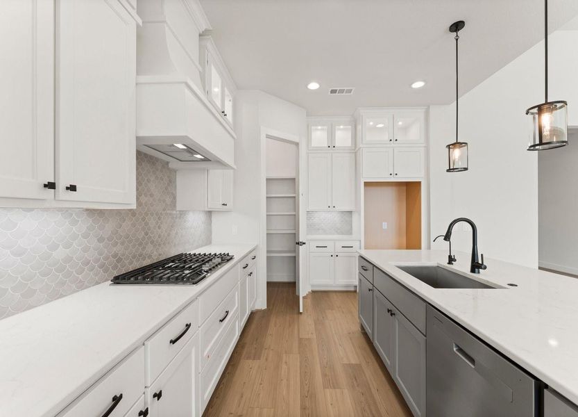 This modern kitchen features sleek white cabinetry, a stylish tile backsplash, and a spacious island. It includes stainless steel appliances, pendant lighting, and a walk-in pantry, all set on elegant hardwood flooring. Perfect for both cooking and entertaining.
