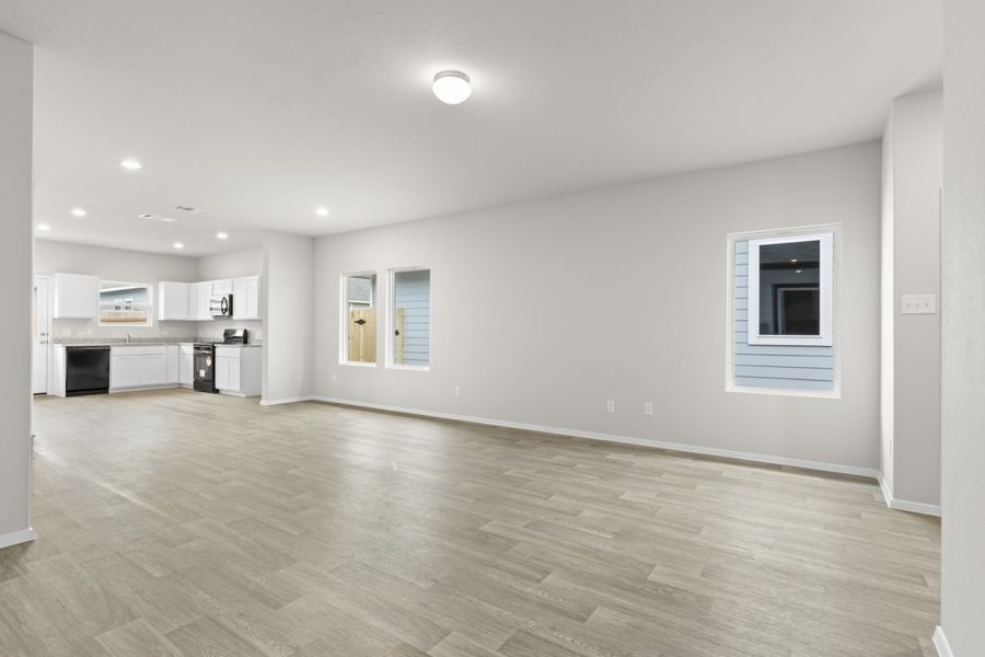 Image of a living room with cream walls, light brown flooring, windows, and a white L-shaped kitchen in the distance