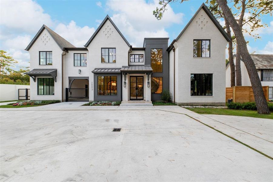 This is a modern, two-story home featuring a mix of white stucco and dark gray accents, large windows, and a striking gabled roofline. The front entrance has a welcoming feel with a covered porch area. The property includes a spacious driveway and a contemporary fence to the side, offering a blend of elegance and privacy.