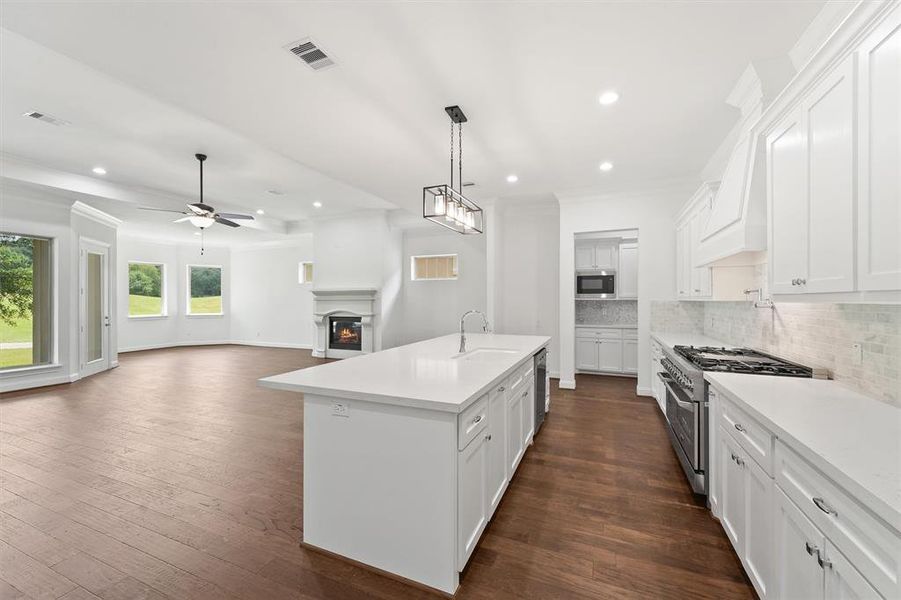 This is a spacious, modern kitchen with white cabinetry and stainless steel appliances, featuring a large central island and hardwood flooring. The open-plan layout includes a bright living area with a fireplace and access to the backyard through glass doors.