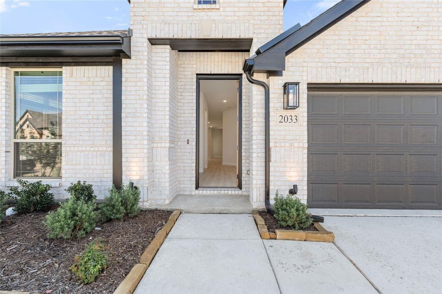 Front entry of home 2033 with white brick exterior, black-framed door, wall sconce, and attached garage Front entry of home 2033 with white brick exterior, black-framed door, wall sconce, and attached garage