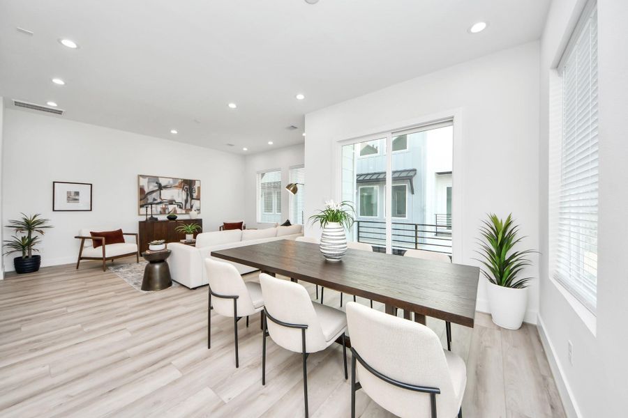 This photo showcases a bright, modern open-plan living and dining area with large windows, light wood flooring, and contemporary furnishings. The space is accented with plants, creating an inviting and airy atmosphere.