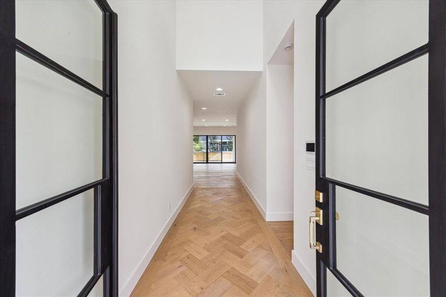 Steel-framed French doors open to a double-height entry with designer chandelier lighting. The space is adorned with engineered white oak flooring in a custom herringbone pattern, designer neutral wall paint, and recessed LED lighting, creating a welcoming and sophisticated ambiance.