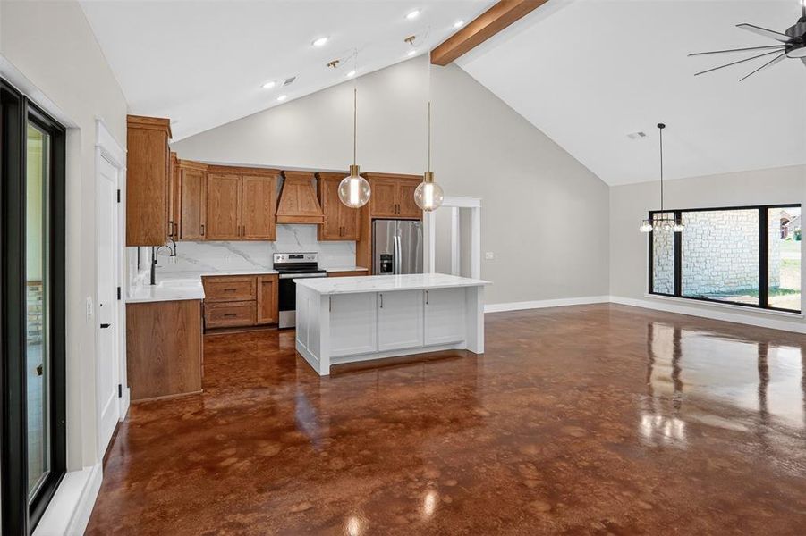 Open concept living area featuring a vaulted ceiling with exposed wooden beam, stained concrete flooring, and a kitchen with wood cabinetry and a central island Open concept living area featuring a vaulted ceiling with exposed wooden beam, stained concrete flooring, and a kitchen with wood cabinetry and a central island
