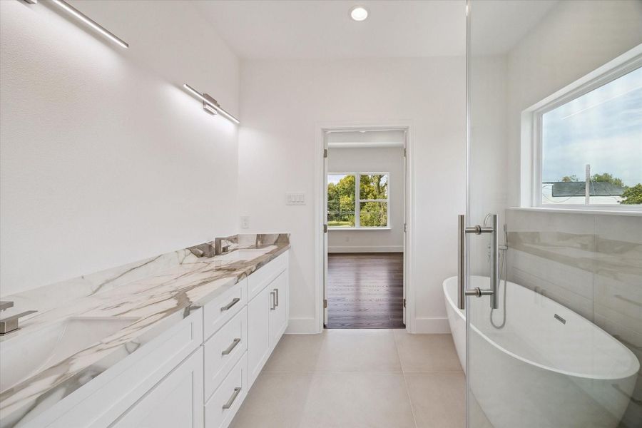 This view of the primary bathroom highlights the generous vanity, dramatic stone-look countertop, sleek modern fixtures, and expansive glass shower enclosure. The freestanding soaking tub adds a luxurious focal point, while large windows introduce abundant natural light throughout the space. Neutral tile flooring and crisp white cabinetry create a bright, polished aesthetic, and the open connection to the primary bedroom reinforces the comfortable, thoughtfully designed suite layout. This view of the primary bathroom highlights the generous vanity, dramatic stone-look countertop, sleek modern fixtures, and expansive glass shower enclosure. The freestanding soaking tub adds a luxurious focal point, while large windows introduce abundant natural light throughout the space. Neutral tile flooring and crisp white cabinetry create a bright, polished aesthetic, and the open connection to the primary bedroom reinforces the comfortable, thoughtfully designed suite layout.