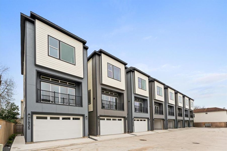 This photo shows a row of modern, multi-story townhomes with a sleek design. Each unit features large windows, a balcony, and an attached garage. The exterior combines light and dark panels for a contemporary look. The area is spacious and well-maintained.