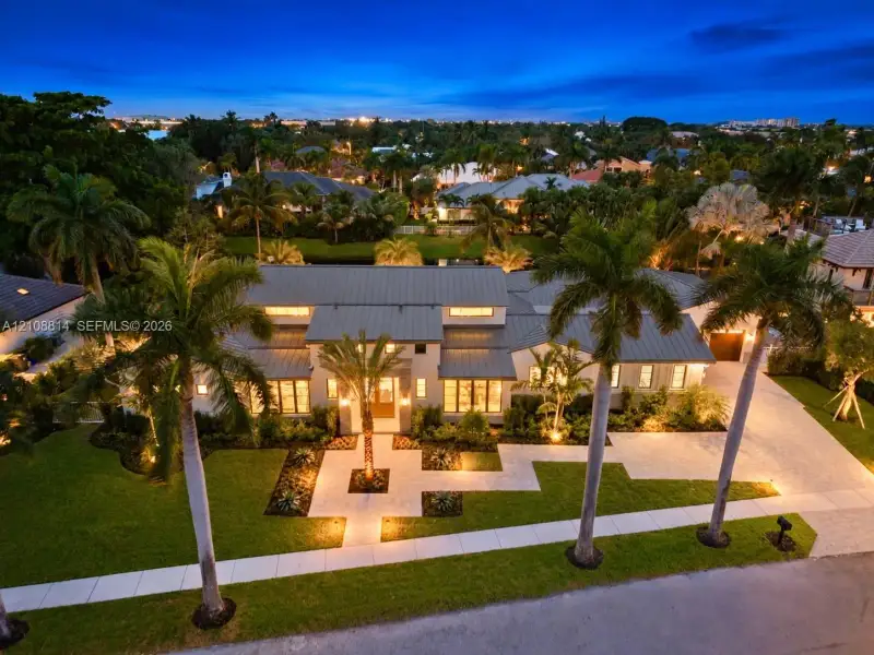 Modern residence featuring a standing-seam metal roof, paver driveway, manicured lawn, stone-surface entryway, and mature palm landscaping Modern residence featuring a standing-seam metal roof, paver driveway, manicured lawn, stone-surface entryway, and mature palm landscaping