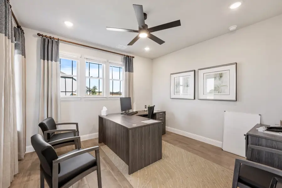 Home office with dark wood L-shaped desk, two guest chairs, ceiling fan, and light hardwood floors