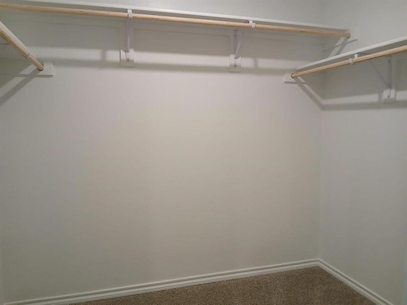 Spacious closet featuring multiple wood closet rods on white shelving brackets, light-colored carpeting, and white baseboard trim Spacious closet featuring multiple wood closet rods on white shelving brackets, light-colored carpeting, and white baseboard trim