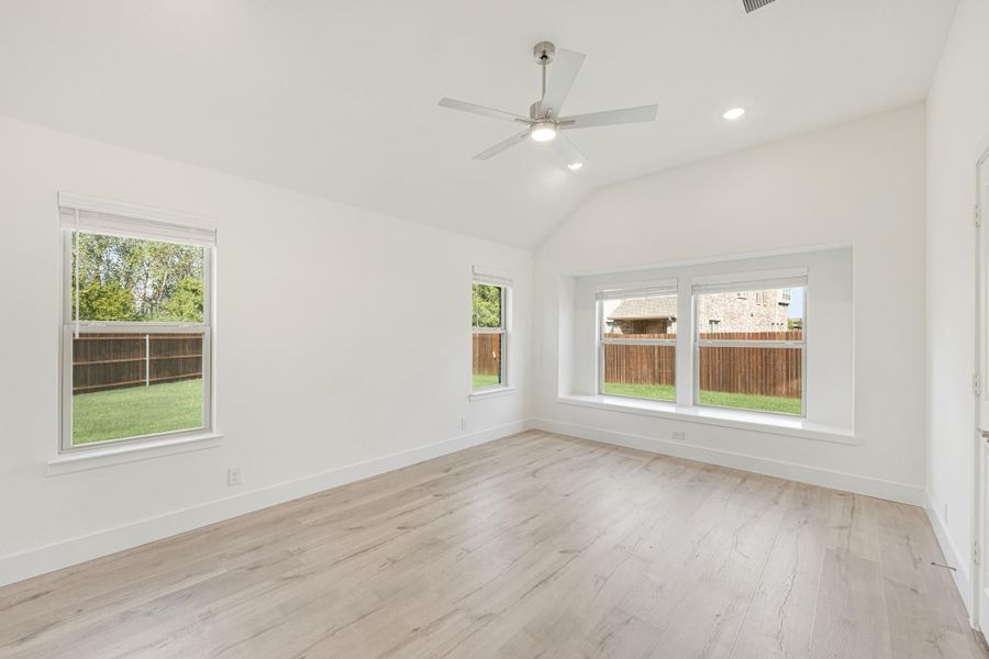 Empty room with vaulted ceiling, ceiling fan, light wood floors, and multiple windows with backyard views