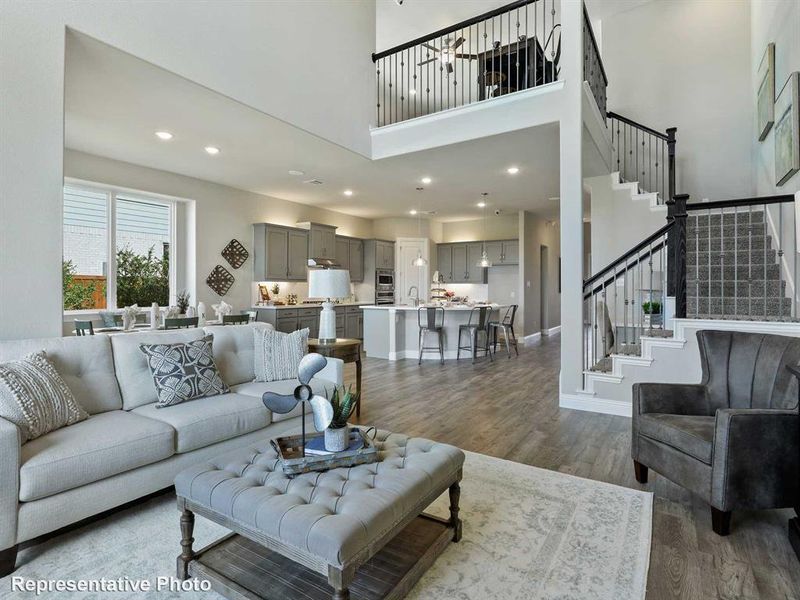 Two-story living area with wood-finish flooring, a grand staircase with wrought iron balusters, a kitchen featuring light gray cabinetry and an island, and an upper-level balcony