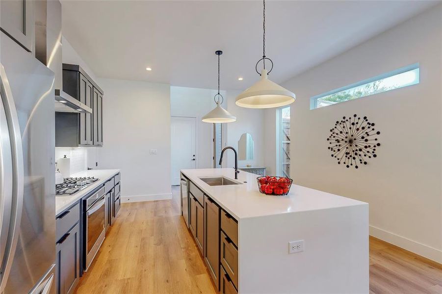 Kitchen with stainless steel appliances, sink, hanging light fixtures, light hardwood / wood-style floors, and an island with sink