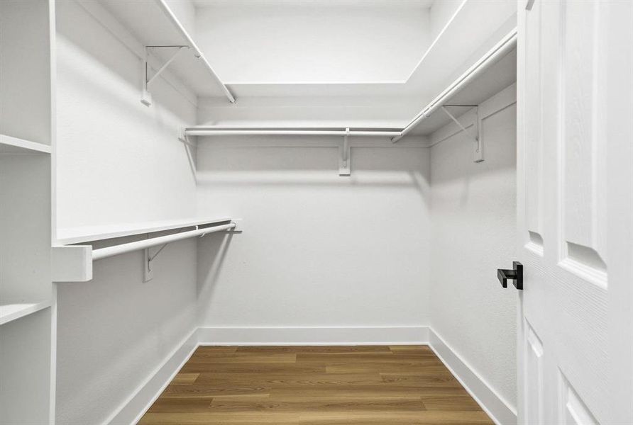 Walk-in closet featuring wood-finish flooring, white built-in shelving, multiple hanging rods, and a white paneled door with matte black hardware Walk-in closet featuring wood-finish flooring, white built-in shelving, multiple hanging rods, and a white paneled door with matte black hardware