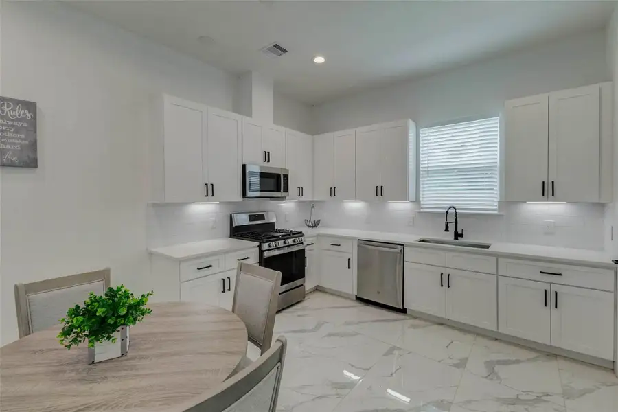 Sleek modern kitchen featuring white shaker-style cabinetry, black hardware, quartz-look counters, subway tile backsplash, stainless gas range, microwave, dishwasher, and under-cabinet lighting. The bright window and glossy marble-look flooring enhance the clean contemporary design.