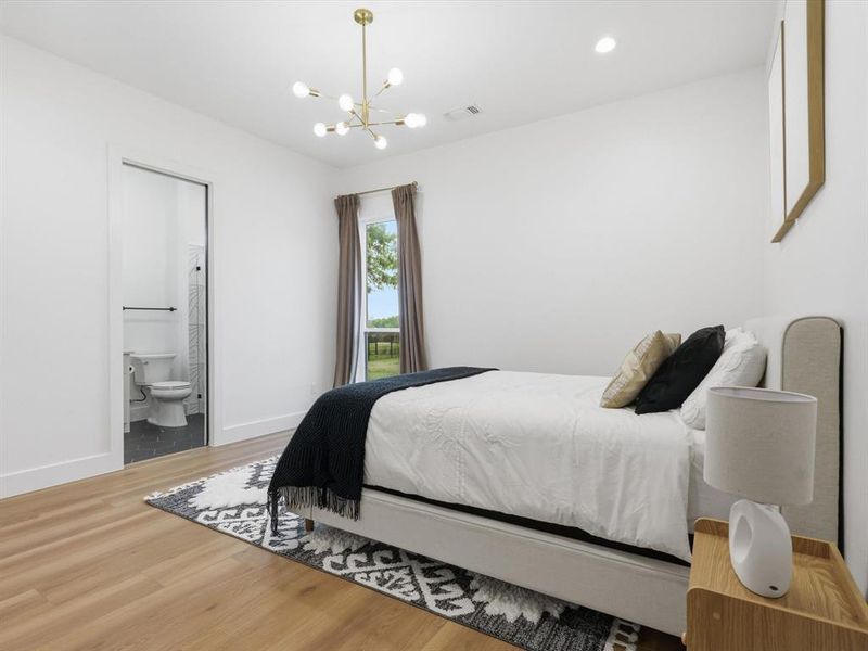 Spacious room featuring wood-finish flooring, a contemporary gold chandelier, a window with outdoor scenery, and an attached private full bath with a pocket door and hexagon tile flooring