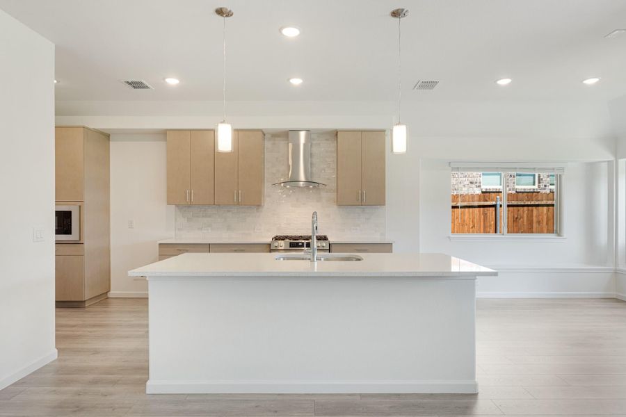 Open kitchen with large white island, light wood cabinets, stainless range hood, and pendant lights over tile backsplash Open kitchen with large white island, light wood cabinets, stainless range hood, and pendant lights over tile backsplash