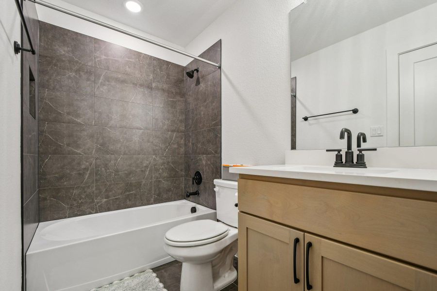 Bathroom featuring a tub and shower with gray tiled surround, a light wood vanity with a white countertop, and modern matte black fixtures Bathroom featuring a tub and shower with gray tiled surround, a light wood vanity with a white countertop, and modern matte black fixtures