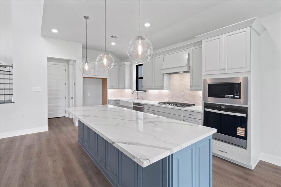 The open-concept gourmet kitchen designed for both cooking and entertaining. It features elegant cabinetry, pristine quartz countertops, and a large central island that doubles as a functional breakfast bar. The cooking area is a chef's dream with a sleek stainless steel gas cooktop, with a wall oven and microwave, all complemented by a chic tile backsplash.