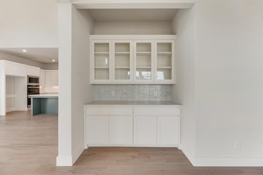 Bar featuring white cabinets, decorative backsplash, light wood-style flooring, glass insert cabinets, and stainless steel microwave Bar featuring white cabinets, decorative backsplash, light wood-style flooring, glass insert cabinets, and stainless steel microwave