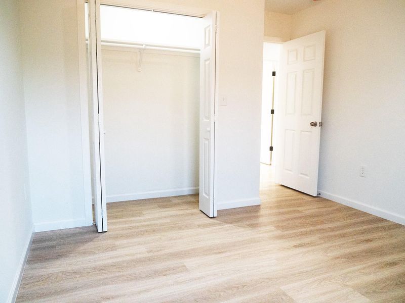 This bright room features light wood flooring and a spacious closet with white bi-fold doors. The open entryway leads to a well-lit hallway, enhancing the airy feel.