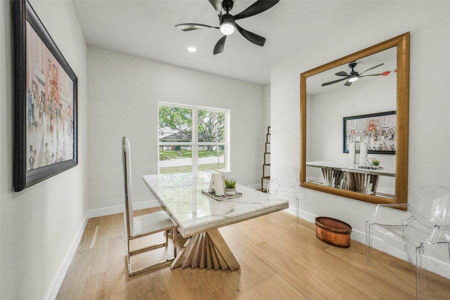 Bright and modern dining area featuring a large window, a sleek glass table with a unique base, and transparent chairs. The room is accented by a large mirror and contemporary art, with light wood flooring and a ceiling fan for comfort.
